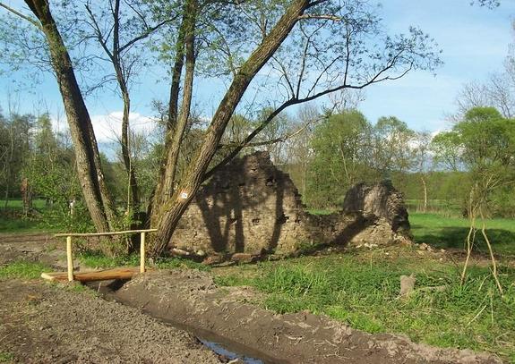 Eine kleine Ruine mit einem kahlem Baum im Hintergrund. Zu sehen ist im Vordergrund ein Bächlein mit einer kleinen Holzbrücke