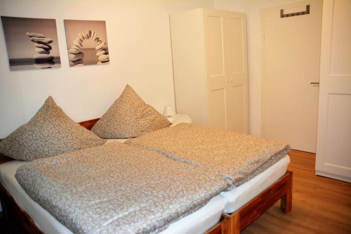 Bild aus dem Schlafzimmer. Bett mit hellgrauer Bettwäsche und an der Wand des Kopfbettes zwei hellgraue Leinwandbilder und rechts ein weißer Kleiderschrank