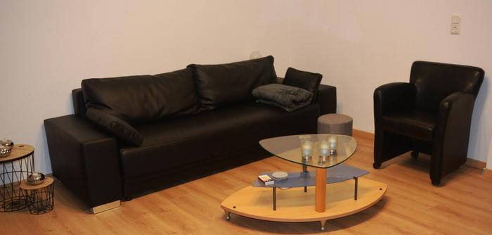Foto vom Wohnzimmer. 3er-Sitz Ledersofa in schwarz und ein Einzelsessel, ebenso aus schwarzem Leder. Holz-Glas-Couchtisch und heller Eichenholzboden