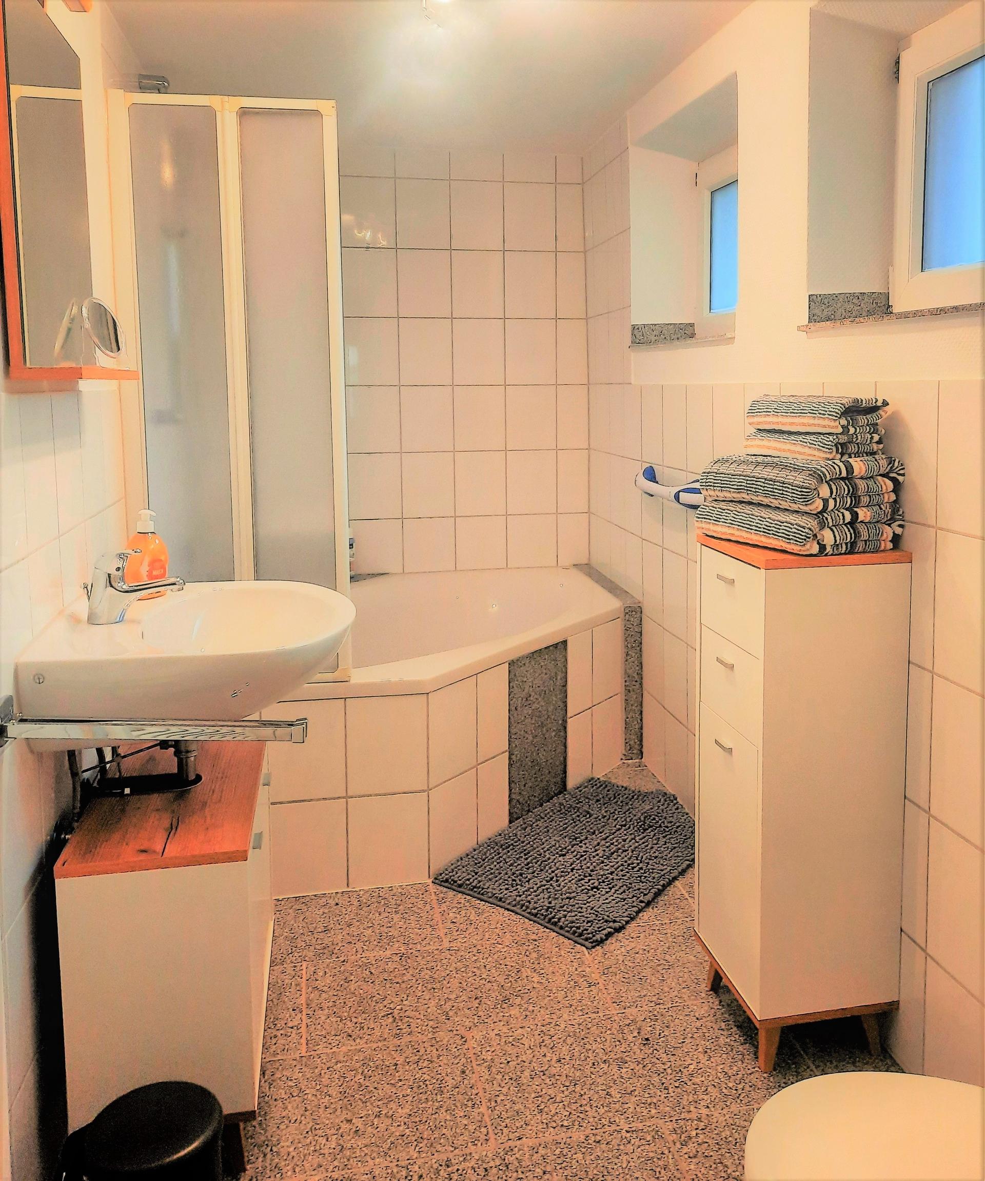 Foto von einem hellen Badezimmer. Es befindet sich eine Eckbadewanne mit einer Glasduschwand.  Rechts befindet sich ein Waschbecken mit Unterschrank und links ein halbhoher Schrank mit gefalteten dunkelblauen Handtüchern.