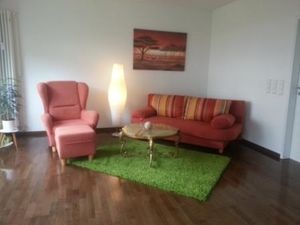 Wohnzimmer.  Roter Sessel und Couch mit einem Couchtisch auf einem grünen Teppich. Der Boden besteht aus braunem Parkett.