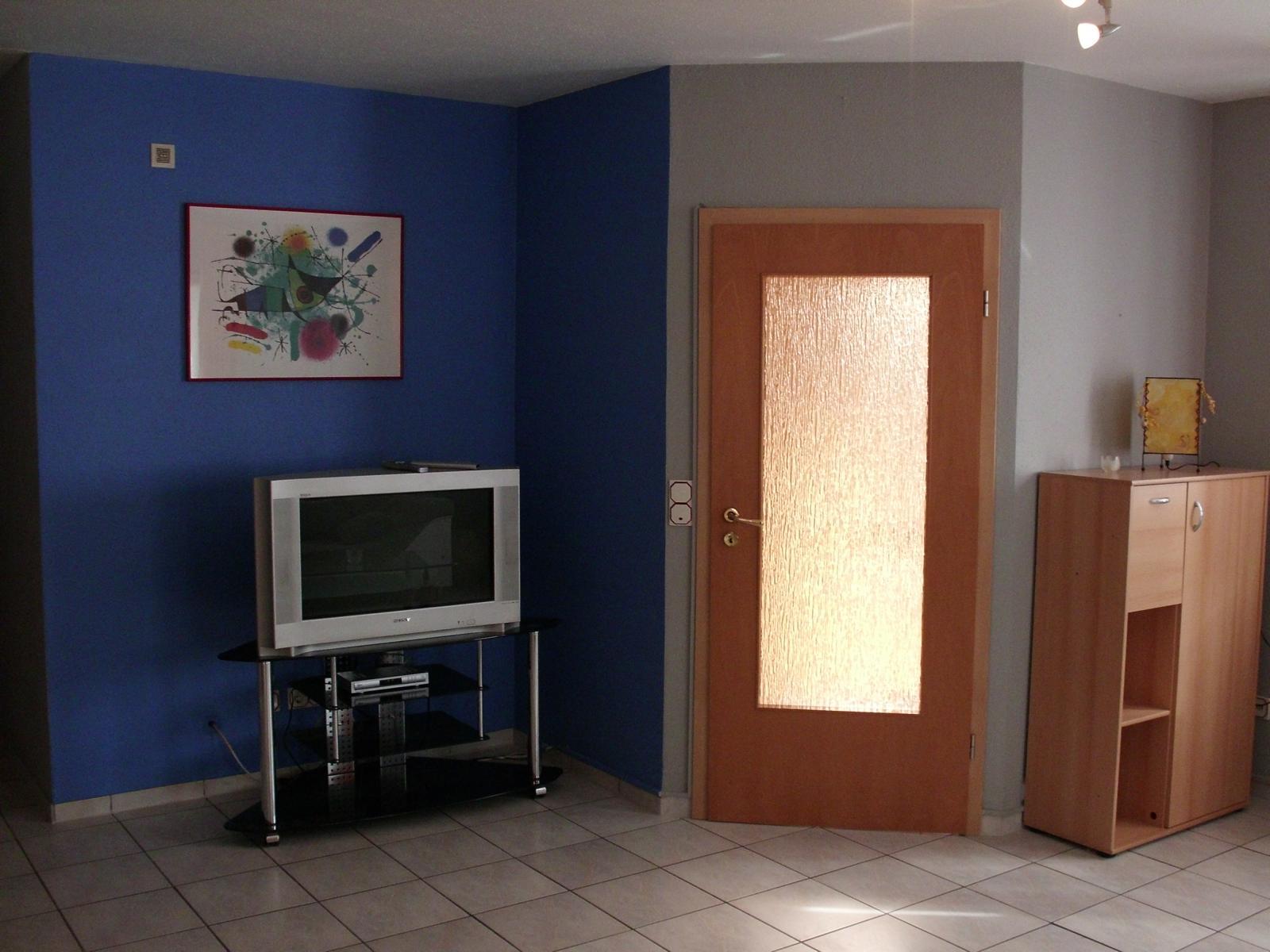 Vor einer lila Wand steht ein Fernseher auf einer Fernseherbank. Rechts daneben ist eine Zimmertür mit Glasausschnitt. 